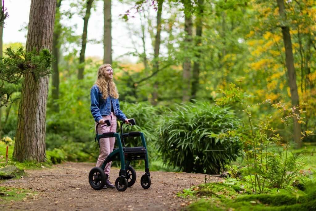 Une jeune femme aux cheveux longs et blonds, à lunettes, se tient debout au milieu de la forêt. Elle est une PMR et se déplace à l'aide d'un déambulateur pour faciliter sa marche.