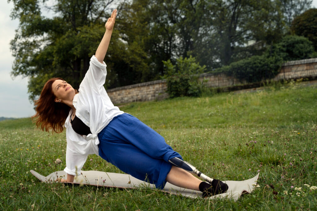 Une jeune femme portant une prothèse à la jambe gauche, fait du yoga en pleine nature. son tapis est posé dans l'herbe. Le yoga est une activité accessible aux personnes handicapées